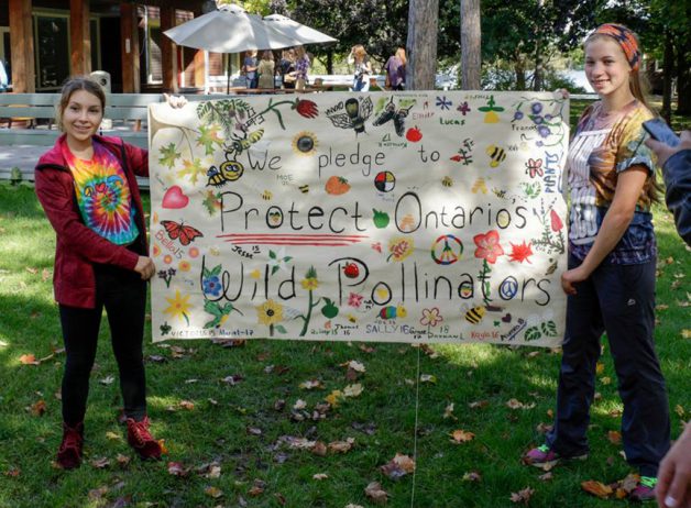 Two girls are outdoors holding up a poster that says: Pledge to Protect Ontario's Wild Pollinators