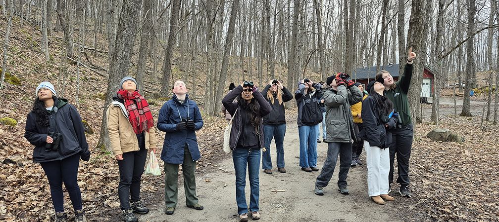 A group of 12 young people enjoy early morning birding at Kilcoo Camp on the 2026 Ontario Nature Youth Council Wetlands Field Trip in a forest near wetlands